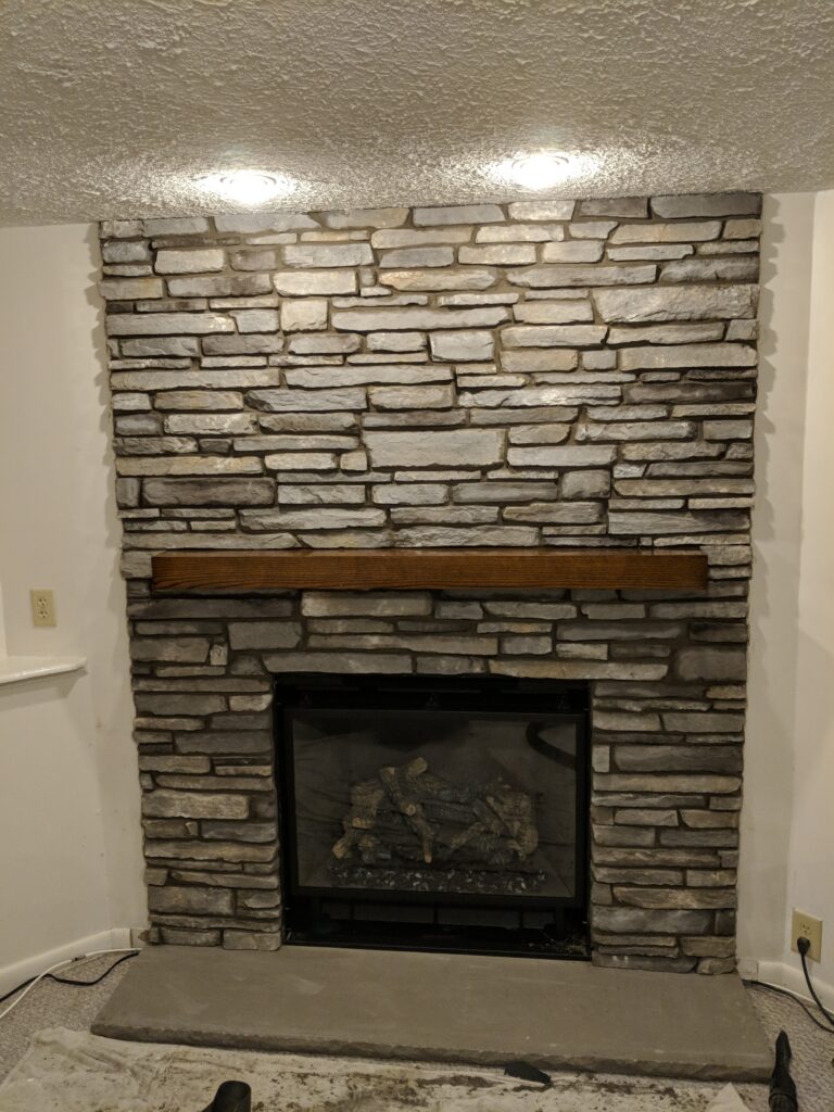 Completed ledgestone stone veneer fireplace surround in light grey and white with wood mantel shelf and recessed lighting, Rochester NY