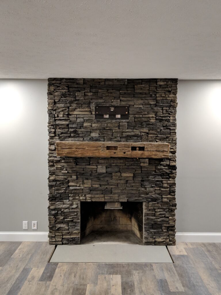 Angled view of dark charcoal ledgestone veneer fireplace surround with rustic beam mantel and recessed ceiling lights, Rochester NY
