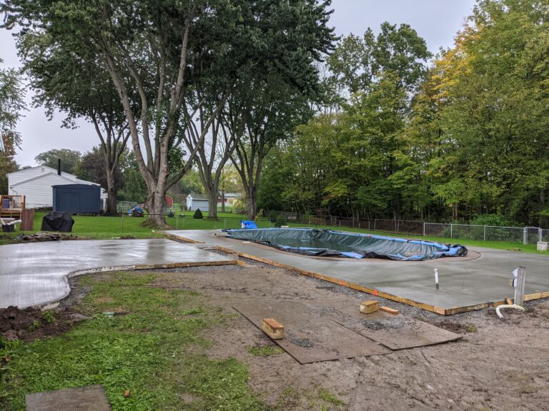 Completed broomed concrete pool deck surrounding a rectangular in-ground pool at a suburban Rochester NY home, photographed at dusk