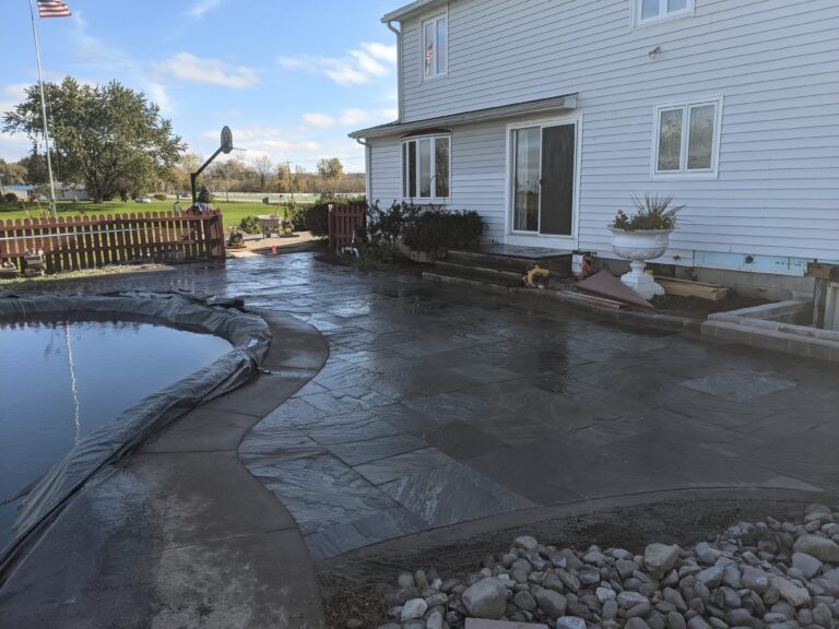 Natural stone tile patio surrounding in-ground pool with stone planter edge beside white home, Rochester NY