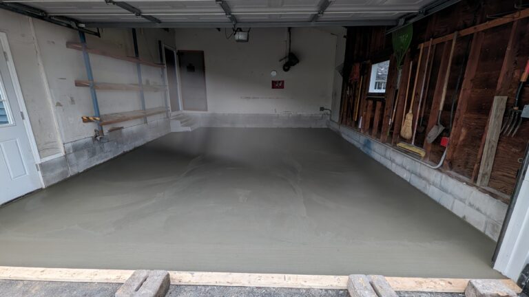 Wide view of freshly poured concrete garage floor with wooden forms and cinder block weights at threshold in Rochester NY