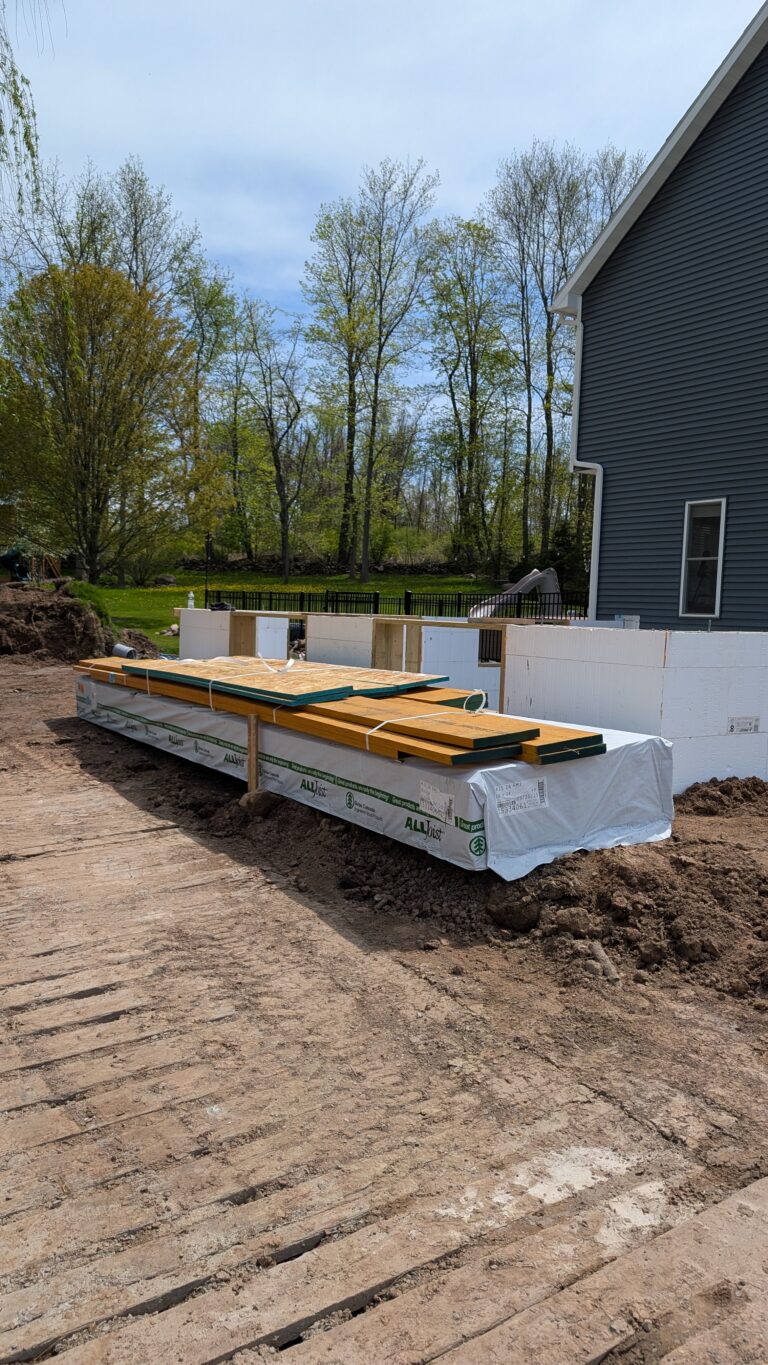 Framing lumber staged on site beside completed poured concrete foundation with waterproofing – Rochester NY