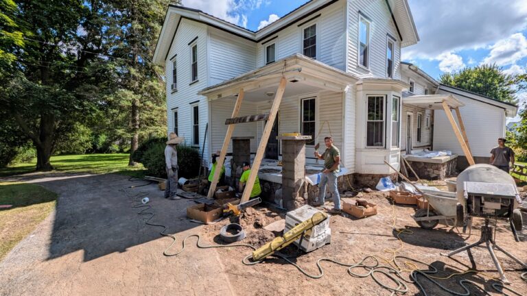 Crew rebuilding front porch stone columns and foundation with temporary bracing on a historic white farmhouse