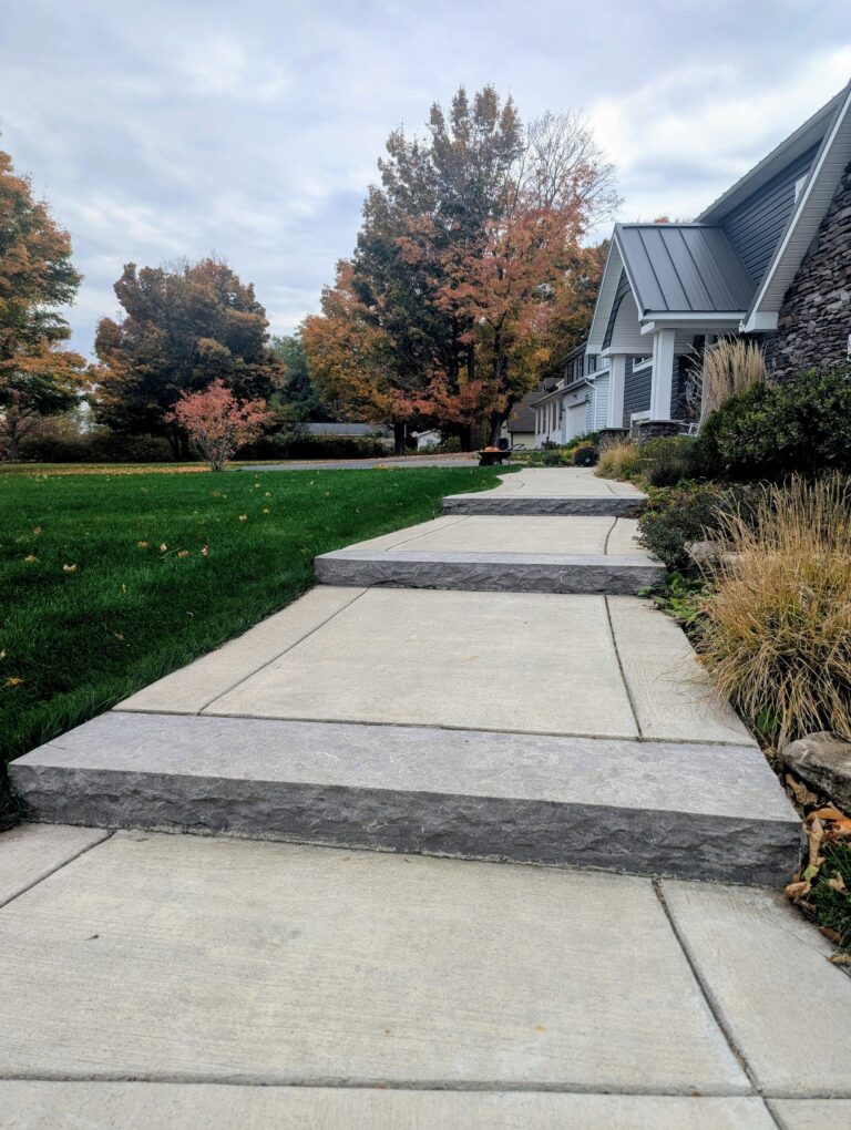 Long broomed concrete walkway with natural-cut stone slab border edges alongside home surrounded by fall foliage, Rochester NY