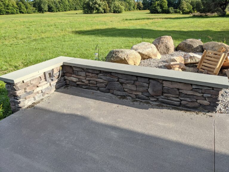 Dry-stack stone veneer seat wall with concrete cap beside gravel patio and natural boulders, Rochester NY