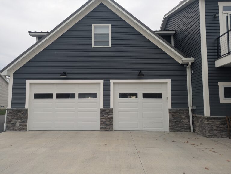 Dark grey ledgestone veneer on residential foundation flanking white garage doors with dark blue siding, Brockport NY