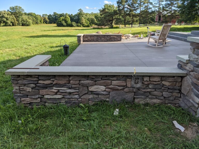 Fieldstone veneer knee wall with concrete cap bordering backyard concrete patio with rocking chair, rural Stafford NY